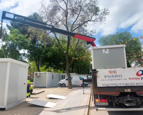 Vista frontal de dos módulos prefabricados tipo camerino instalados por Europa Prefabri para el Ayuntamiento de Pinto, con acceso independiente y acabados en blanco. En el entorno se aprecia una zona urbana destinada a actividades culturales.