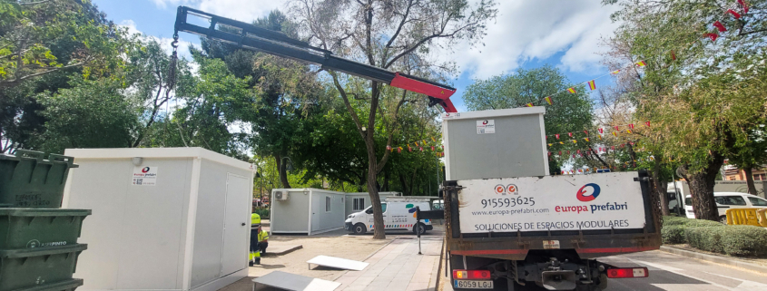 Vista frontal de dos módulos prefabricados tipo camerino instalados por Europa Prefabri para el Ayuntamiento de Pinto, con acceso independiente y acabados en blanco. En el entorno se aprecia una zona urbana destinada a actividades culturales.